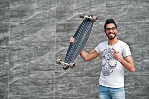 homem árabe de estilo de rua em óculos com longboard posou contra a parede cinza. dedos de pedra mostram. foto