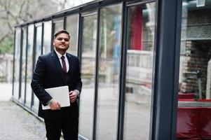 empresário indiano elegante com roupa formal com laptop nas mãos de pé contra janelas no centro de negócios. foto