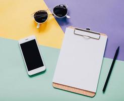 mesa de trabalho criativa de estilo plano leigo com smartphone, prancheta em branco e óculos de sol em fundo colorido moderno foto