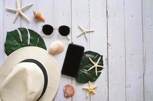 plano de conceito de itens de verão, chapéu, óculos de sol, smartphone e concha no fundo da mesa de madeira branca com espaço de cópia, vista superior foto