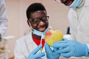 médico masculino americano africano em clínica odontológica com uma maçã e escova de dentes nas mãos. foto