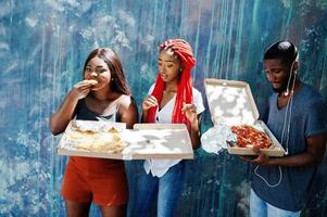 três alegres amigos afro-americanos segurando caixas de pizza contra a parede de pintura. foto