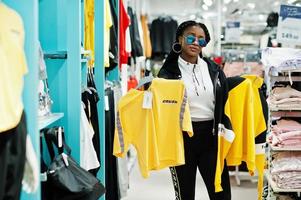 mulheres afro-americanas em agasalhos e óculos de sol, compras no shopping de roupas esportivas contra prateleiras. ela escolhe camiseta amarela. tema da loja de esportes. foto