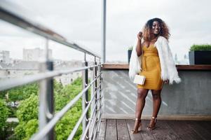 glamour mulher afro-americana de vestido amarelo e capa de lã branca com bolsa posada na varanda. foto