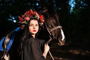 garota mística em grinalda veste em preto com cavalo em madeira. foto