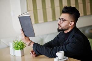 homem árabe usar jaqueta jeans preta e óculos sentado no café, ler o livro e beber café. cara modelo árabe elegante e elegante. foto