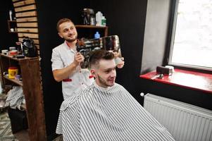 jovem barbudo cortando o cabelo pelo cabeleireiro enquanto está sentado na cadeira na barbearia. alma de barbeiro. foto