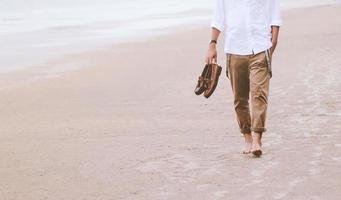 homem sozinho andando na praia carregando sapatos de couro foto