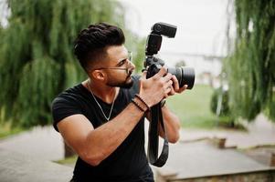 Fotógrafo de homem macho de barba árabe linda e bonita em óculos e camiseta preta com câmera profissional nas mãos. foto