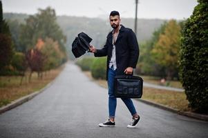 homem de barba árabe alto na moda usa casaco preto com guarda-chuva e bolsa posada no dia de chuva. foto