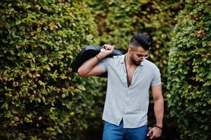 homem de barba árabe alto na moda usar camisa, jeans e óculos de sol posou no parque e segure a mala. foto