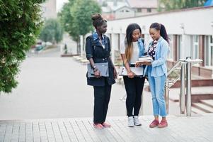 três estudantes africanos femininos posaram com mochilas e itens escolares no pátio da universidade e olham para o tablet. foto