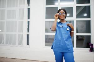 retrato de feliz feminino americano africano jovem médico pediatra no casaco uniforme azul e estetoscópio contra a janela no hospital. saúde, médico, especialista em medicina - conceito. foto