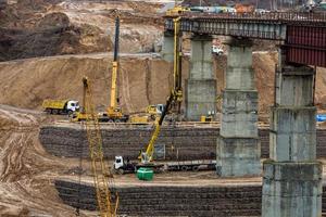 construção ou reconstrução de ponte de concreto rodoviário sobre um rio largo. máquinas de construção, caminhões e guindastes trabalham foto