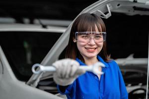 conceito de reparo automático. jovem mecânico asiático de uniforme detém o conceito de chave e reparo. serviço e manutenção de automóveis. uma mulher mecânica com uma prancheta verifica o motor de um carro na garagem. foto