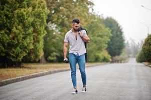 homem de barba árabe alto na moda usar camisa, jeans e óculos de sol andando no parque com guarda-chuva e casaco na mão. foto