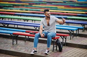 homem de barba árabe alto na moda usar camisa, jeans e óculos de sol, sentado na fileira colorida de bancos e usar seu telefone. foto