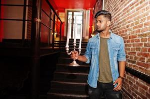 retrato de bonito bem sucedido barbudo sul-asiático, jovem indiano freelancer em camisa jeans azul em pé na boate e segure o candelabro. foto