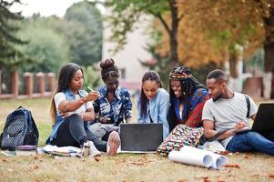 grupo de cinco estudantes universitários africanos passando tempo juntos no campus no pátio da universidade. amigos negros afro sentados na grama e estudando com laptops. foto