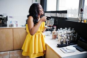 elegante mulher afro-americana em vestido amarelo posou na cafeteria com uma xícara de cappuccino. foto