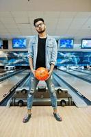 homem asiático de barba elegante na jaqueta jeans e óculos em pé na pista de boliche com bola na mão. foto