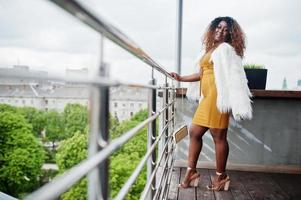 glamour mulher afro-americana de vestido amarelo e capa de lã branca com bolsa posada na varanda. foto