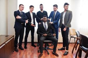 seis homens de negócios multirraciais em pé no escritório e rolar o homem na cadeira. grupo diversificado de funcionários do sexo masculino com roupa formal se divertindo. foto