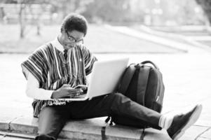 estudante de homem africano em roupas tradicionais e óculos com mochila, trabalhando no laptop com celular ao ar livre. foto