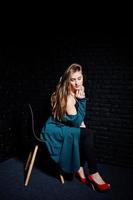 menina morena bonita usa jaqueta preta e verde de salto alto vermelho, sentado e posando na cadeira no estúdio contra a parede de tijolos escuros. retrato de modelo de estúdio. foto