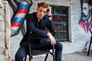 jovem macho macho elegante na jaqueta preta posou ao ar livre da rua. homem modelo incrível sentado contra a parede de grafite da barbearia. foto
