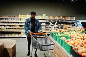 homem afro-americano casual elegante na jaqueta jeans e boina preta na seção orgânica do supermercado com carrinho de compras. foto