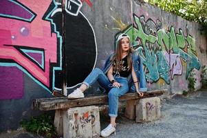 menina elegante casual hipster em boné e jeans usa música de fones de ouvido do telefone celular contra grande parede de graffiti com bomba. foto