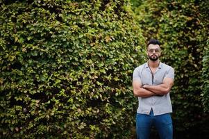 homem de barba árabe alto na moda usar camisa, jeans e óculos de sol posou no parque contra vegetação. foto
