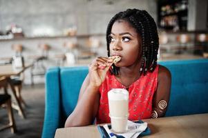 mulher afro-americana atraente sentada à mesa no café com café com leite e comendo biscoito. foto