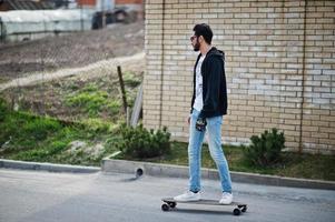 homem árabe de estilo de rua em óculos com longboard longboard na estrada. foto