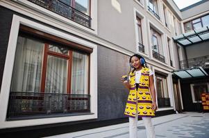 mulheres afro-americanas elegantes na jaqueta amarela posaram e fones de ouvido na rua contra o edifício moderno com bebida quente em copo de papel descartável. foto