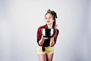 jovem dona de casa engraçada na camisa quadriculada e short amarelo pin up estilo com panela e colher de cozinha isolado no fundo branco. foto