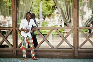 homem afro-americano elegante na camisa branca e calça colorida com chapéu posou ao ar livre. menino modelo preto na moda. foto