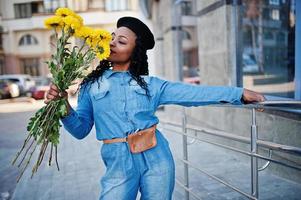 elegantes mulheres afro-americanas em jeans usam e boina preta com buquê de flores amarelas posadas ao ar livre em dia ensolarado contra edifício moderno azul. foto