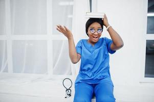 retrato de feliz feminino americano africano jovem médico pediatra no casaco uniforme azul e estetoscópio com livros nas mãos. saúde, médico, especialista em medicina - conceito. foto