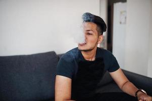 homem indiano bonito e elegante de preto sentado no quarto, fumando narguilé foto