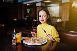 menina morena engraçada de suéter amarelo comendo pizza no restaurante. foto