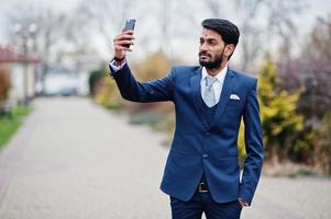 homem de negócios indiano de barba elegante com bindi na testa, usar terno azul posou ao ar livre fazendo selfie no celular. foto