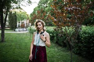 menina modelo loira elegante e bonita na saia de veludo vermelho elegante, blusa branca e chapéu, sentado na grama verde no parque com telefone e fones de ouvido. foto