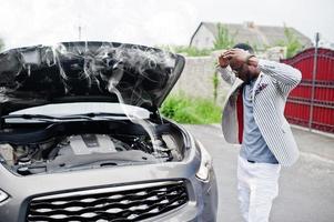 homem afro-americano elegante e rico fica na frente de um carro suv quebrado precisa de assistência olhando sob o capô aberto com fumaça. foto