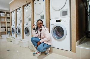 alegre mulher afro-americana em óculos de sol perto da máquina de lavar roupa na lavanderia self-service.