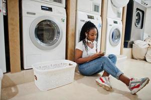 alegre mulher afro-americana sentada com fones de ouvido e lendo revista perto da máquina de lavar roupa na lavanderia self-service. foto