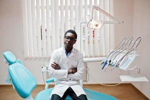 médico masculino americano africano em óculos, sentado na cadeira do dentista na clínica odontológica. foto