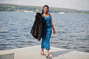 modelo plus size de pele escura afro-americana posou em um vestido azul brilhante e casaco de pele preto contra o lado do mar. foto