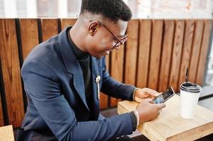 homem afro-americano incrivelmente bonito usa blazer azul com broche, gola alta preta e óculos posados na rua. cara preto elegante com xícara de café olhando para o telefone dele. foto